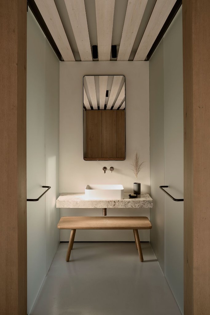 The bathroom is minimalist, with a stone vanity, wooden bench and frosted glass clad showers