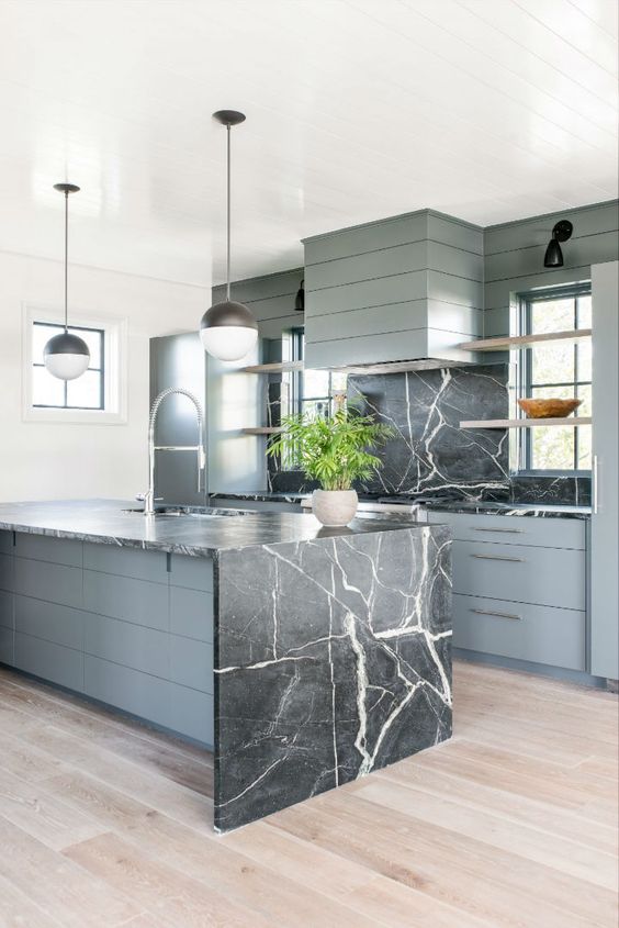 a grey wooden kitchen with a black marble backsplash and a matching island with a waterfall countertop
