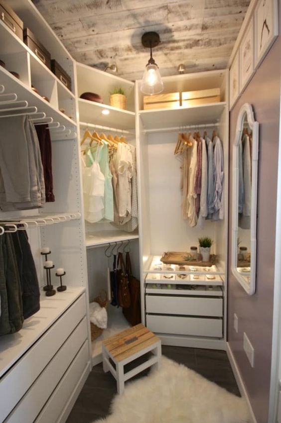 A small farmhouse closet with holders for hangers, shelves with boxes, built in drawers, a wooden stool and a whitewashed ceiling