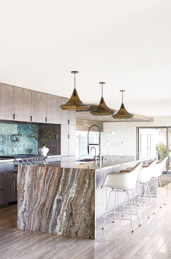 a neutral wooden kitchen with a large kitchen island topped with a gorgeous stone waterflal countertop that brings a luxurious feel