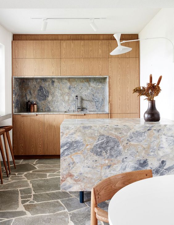 A small light colored kitchen with a grey marble backsplash and countertops including a waterfall one that rocks