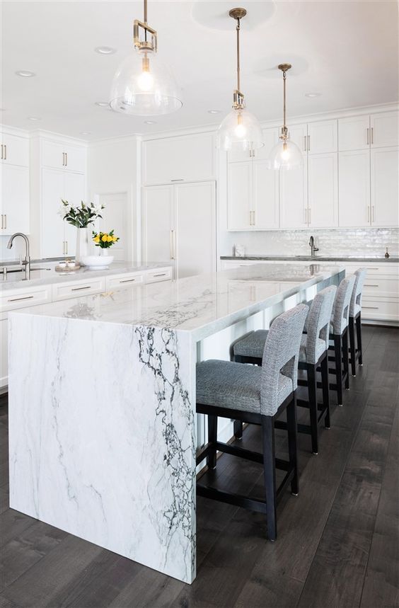 an elegant white kitchen with a chic kitchen island with a white marble waterfall countertop that brings luxe here