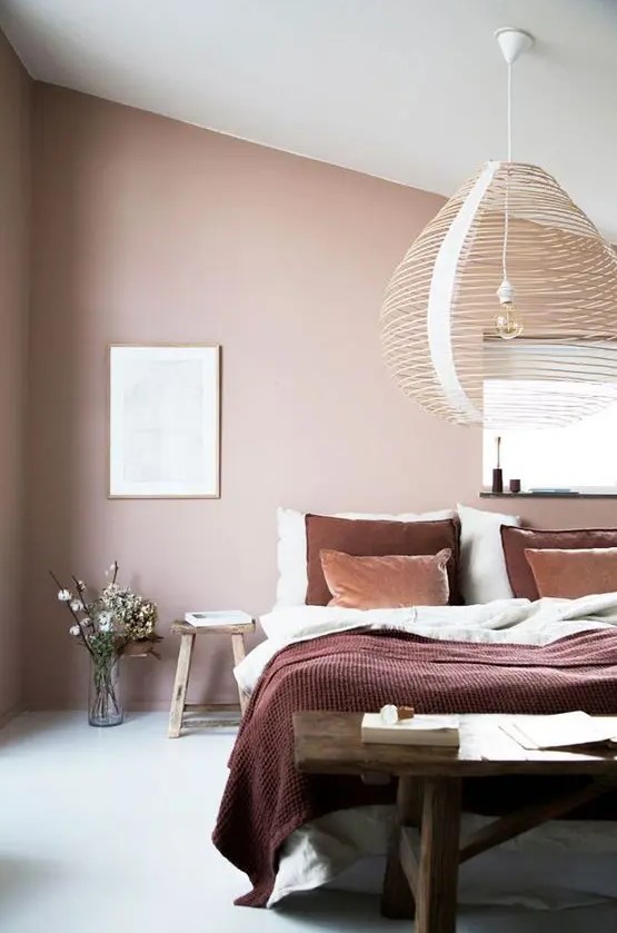 A beautiful attic bedroom with blush walls, a bed with contrasting bedding, a dark stained wooden bench and a woven pendant lamp