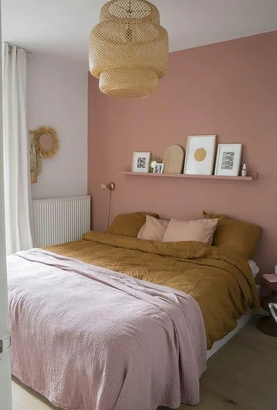 a boho bedroom with a dusty pink accent wall, a bed and a shelf over it, a woven pendant lamp and some pretty pink bedding is chic