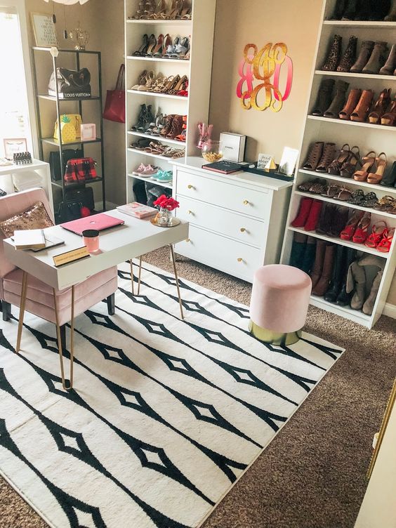 a bold glam home office with layered rugs, a small desk, a pink chair and ottoman, shoe and bag racks