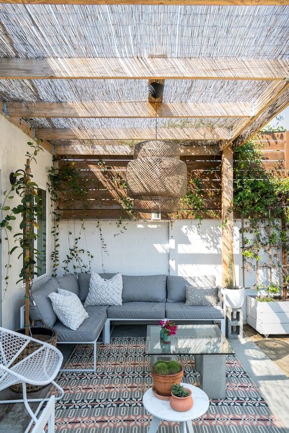 a bright boho patio with a corner sofa, some metal chairs, a concrete and marble table, a wicker pendant lamp and some greenery on the walls