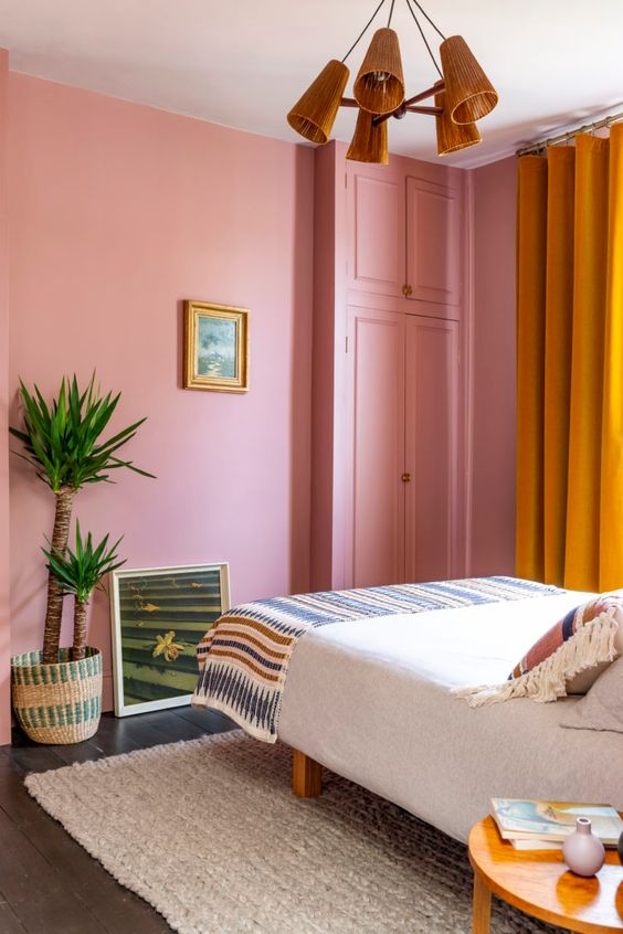 a bright pink bedroom with a bed and printed bedding, yellow curtains, potted plants, a nightstand and a chandelier