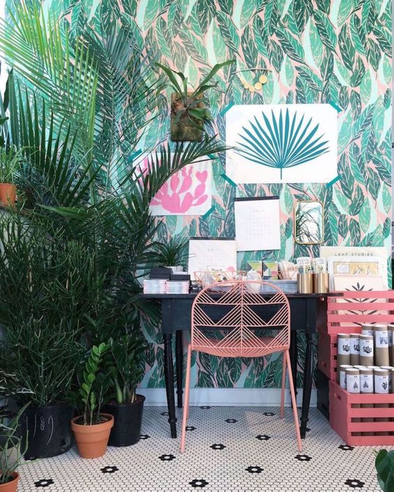 a bright tropical home office with tropical wallpaper, bright artworks and potted plants, a vintage desk and pink furniture