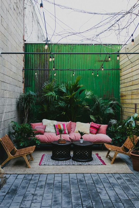a bright tropical patio with a pink sofa and printed pillows, leather chairs, lots of lush plants in pots and black tables