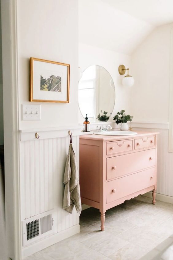 a chic and cute bathroom with white beadboard, a pink vanity, a round mirror, potted greenery and sconces is ethereal