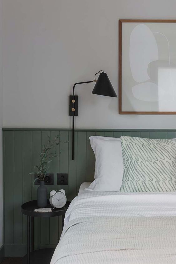 a chic bedroom with green beadboard, a black nightstand and sconce, a neutral artwork and some accessories