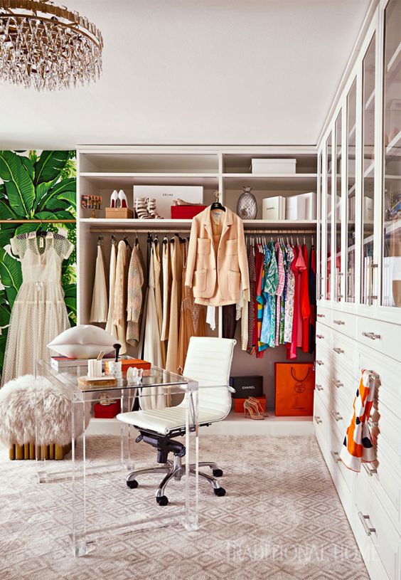 a colorful glam cloffice with a crystal chandelier, a printed rug, a makeshift closet, a largre closed storage unit, an acrylic desk and a white chair