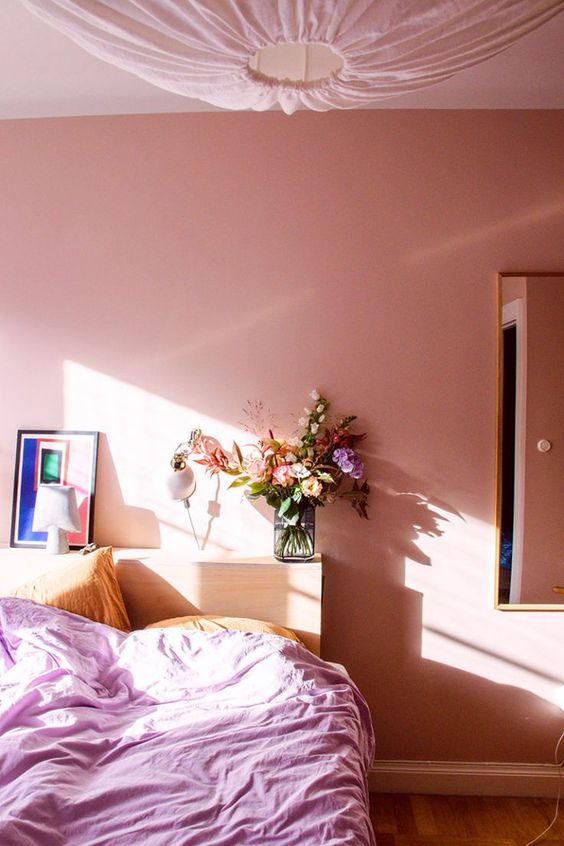 a modern pink bedroom with a bed and a storage headboard, pink and yellow bedding, some bright art and blooms