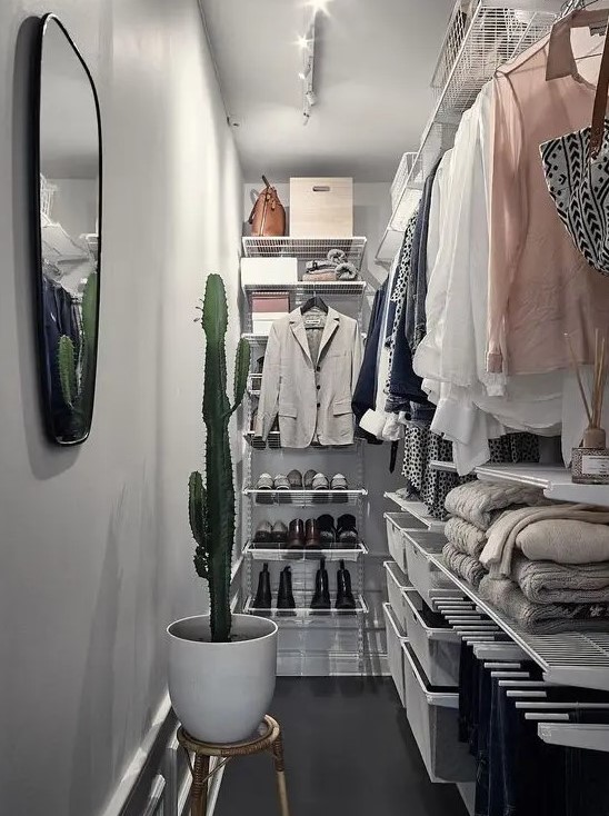 A narrow walk in closet with white airy shelves and baskets for storage, railing and drawers, a mirror and a potted cactus