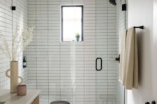 a peaceful and welcoming small bathroom clad with white skinny tiles, with a chic vanity, black fixtures and a small window in the shower