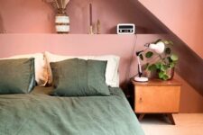 a pink attic bedroom with a built-in shelf with decor, a bed with green bedding, a stained nightstand and greenery