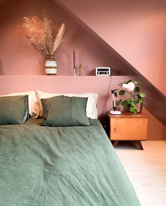 A pink attic bedroom with a built in shelf with decor, a bed with green bedding, a stained nightstand and greenery