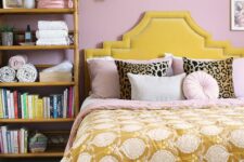 a pink bedroom with a yellow upholstered bed and pink and yellow bedding, a rattan shelf with books and stuff and a gallery wall