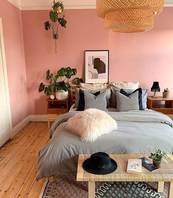 a pink boho bedroom with a bed and grey bedding, a rattan bench, stained nightstands and potted plants