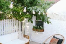 a small and cute patio with a white sofa, wicker chairs, a rattan pouf and string lights over the space
