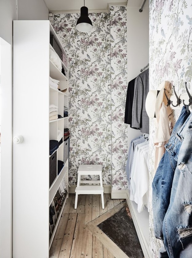 a small and cute walk-in closet with botanical wallpaper, an open wardrobe and racks for hanging clothes plus a stool and a lamp