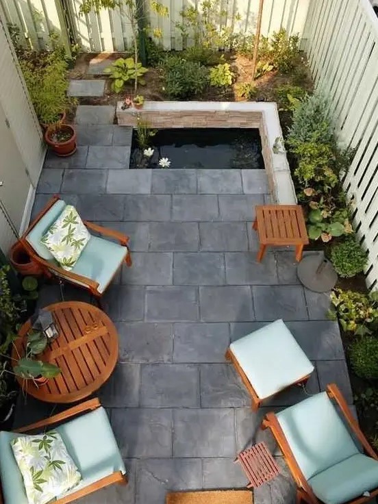 A small and zen like paved patio with tiles on the ground, a tiny pond, some growing plants and cool garden furniture with blue upholstery