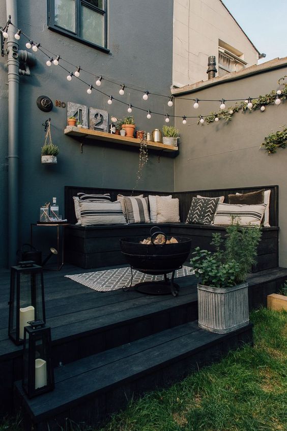 A small black patio with a built in sofa, potted greenery, lights over the space and printed pillows plus a fire pit