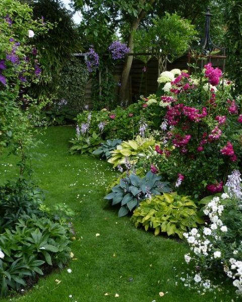 a small colorful garden with a grene lawn, some shrubs, bright blooms, several trees in the corner