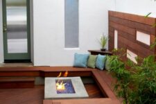 a small contemporary patio clad with wood, with a built-in wooden bench, a fire pit, some potted greenery and pillows