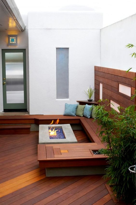 A small contemporary patio clad with wood, with a built in wooden bench, a fire pit, some potted greenery and pillows