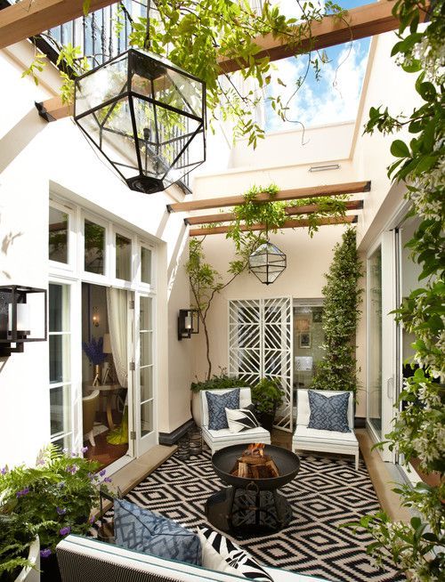 a small contemporary patio in a monochromatic color scheme, with a printed rug, blue pillows, lots of greenery, catchy lanterns and a fire pit
