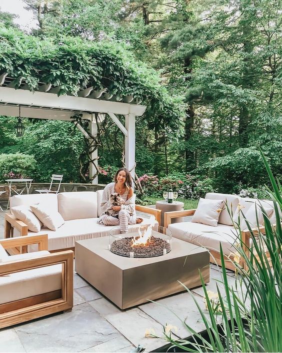 a small contemporary patio with simple neutral furniture, a large fire pit and some greenery and blooms around