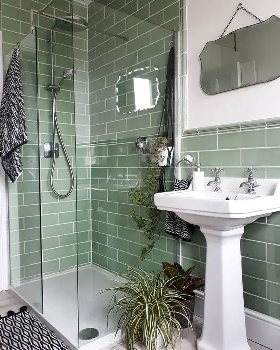 A small green bathroom clad with green subway tiles, with a shower, a free standing sink and a mirror of a catchy shape
