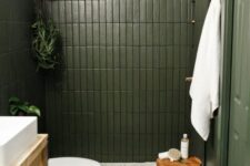 a small green bathroom with skinny tiles, a cane vanity, a wooden stool and some plants and white towels