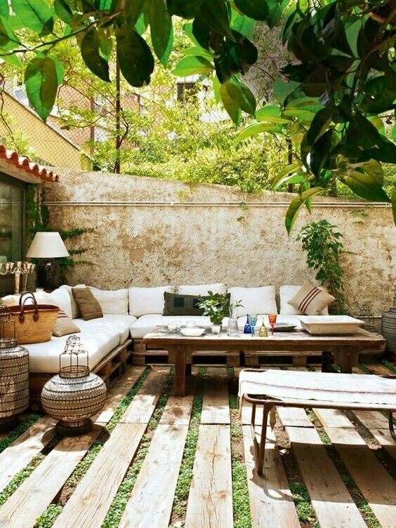 a small patio with a corner sofa, a wooden table, wicker candle lanterns, a wooden plank floor and some greenery