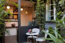 a small patio with a couple of chairs, a small side table, potted greenery, greenery climbing up the wall and some lights and lamps