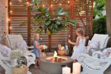 a small patio with a tile floor, a concrete fire pit, vintage chairs, a potted tree, lights, candles and blooms