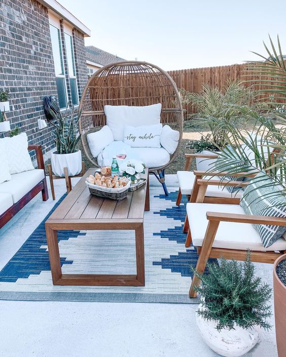 A small patio with white seating furniture, a wooden coffee table, an egg shaped chair, some potted greenery and plants
