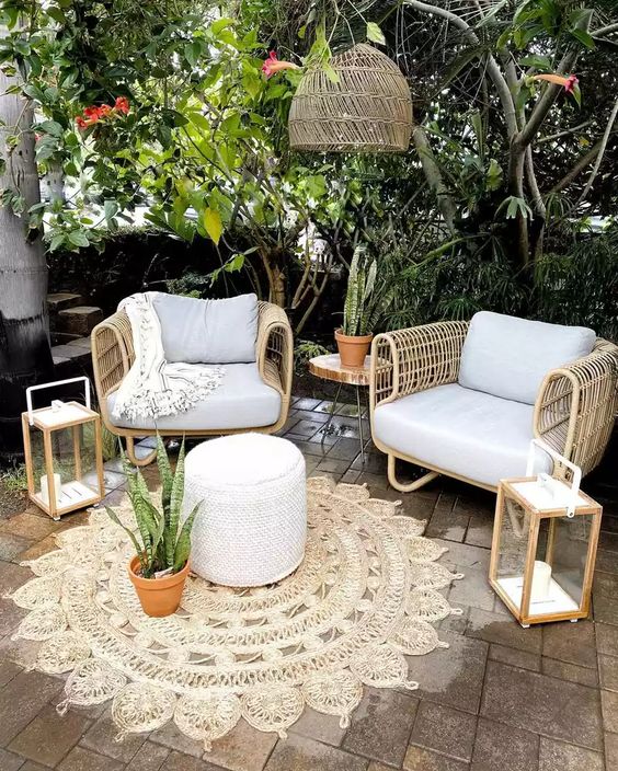 a small patio with wicker furniture, a pouf, some candle lanterns, a woven pendant lamp and greenery around