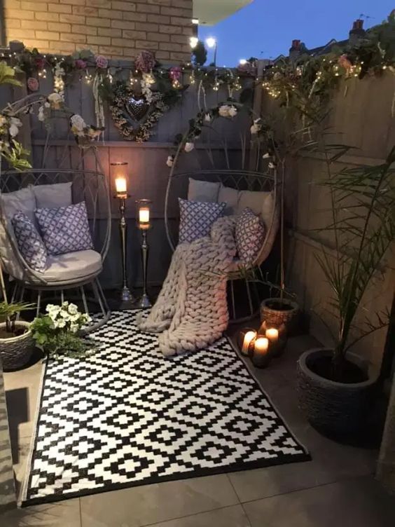 A small patio with egg shaped chairs with pillows, printed pillows and a rug, potted plants and blooms, lights and candles