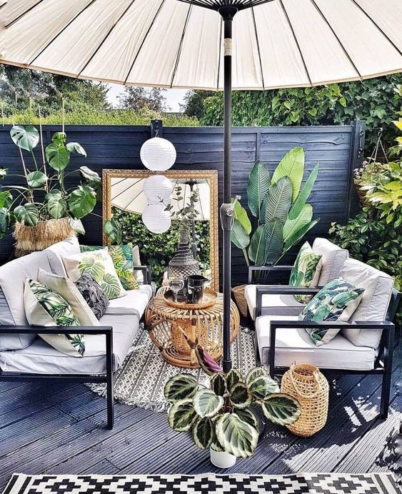 a small tropical patio done in black and grey, with white furniture, a large umbrella, potted plants and tropical pillows