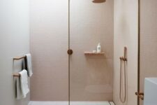 a small unique bathroom clad with blush mini tiles, with neutral and pink ones on the floor and a skylight over the shower plus brass touches