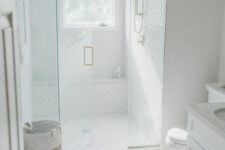 a small white bathroom with large scale and fish scale tiles, a shower space, a white vanity and a basket plus gold fixtures