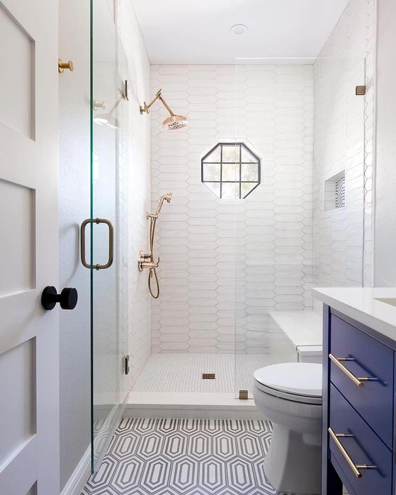 a small yet stylish bathroom with catchy long tiles in the shower and patterned ones ont he floor, with a bold blue vanity, brass touches and a geometric window