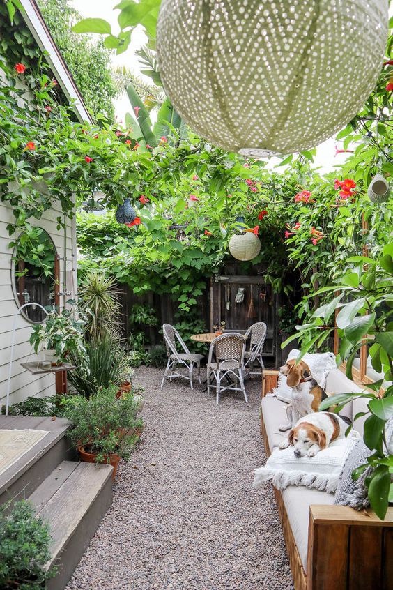 a small yet very lush patio with a sofa, pendant lamps, lots of greeneyr and pink blooms and a small dining set
