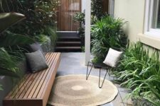 a tiny patio with a wooden bench, growing greenery and shrubs, a black chair, a jute rug is welcoming and cozy