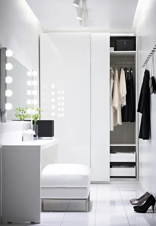a white minimalist closet with a built-in wardrobe with sliding doors and drawers, a vanity with lights and a pouf for sitting