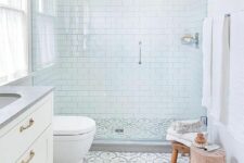 an airy neutral bathroom with white subway tiles and printed ones on the floor, a white vanity, a stool and a tree stump
