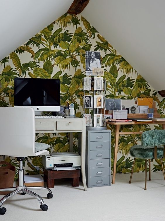 an attic tropical home office with an accent wall, two desks, a green fur chair, a stand with cards and a white chair