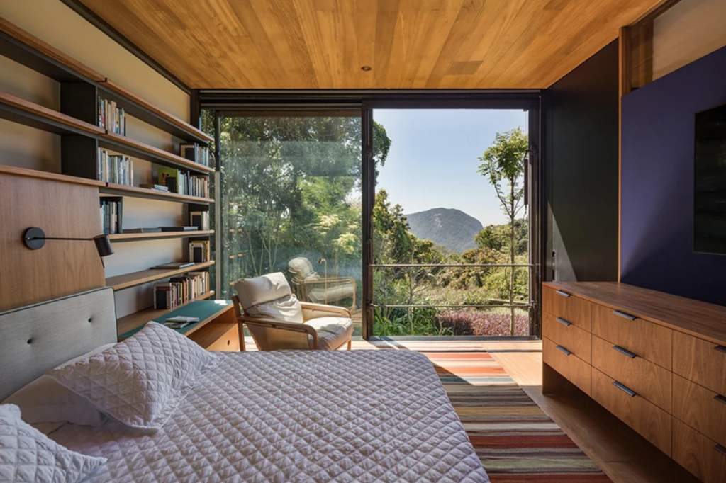The bedroom features a glazed wall, a shelving unit on one wall and a large TV